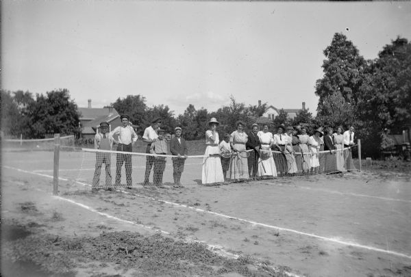 Group portrait of a line of people, some holding tennis rackets, posed in a line behind a net on a dirt tennis court between North 3rd and 4th Street. Houses and trees are in the background.