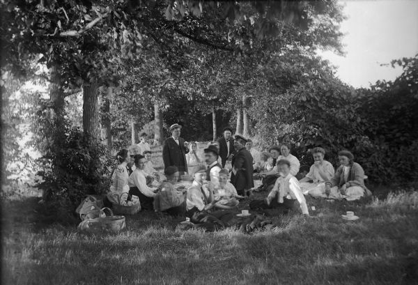 Large group of people at a picnic under the trees at Lasley's Point along Lake Winneconne. Many individuals are in the process of turning toward the photographer, causing their faces to look blurry. Picnic baskets and tea cups lie in the grass around the picnickers, who are gathered around a large cloth set with the food.