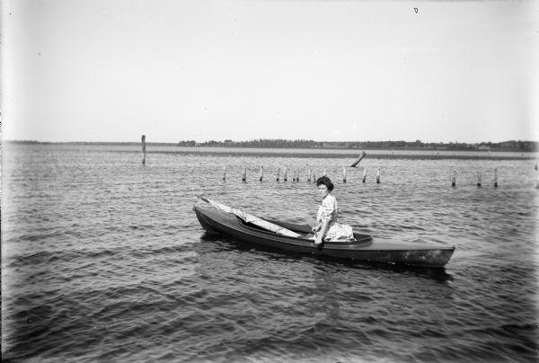 A young woman sits in a small kayak-like boat on Lake Winneconne. A sail wrapped around a mast is laying in front of her on the gunwale. Wooden pilings are protruding from the water behind her. The far shoreline with houses and buildings is in the background.