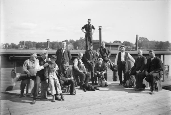 Group of twelve men and one boy posing on a dock. A dog lays at the feet of the man in the center. Three of the men hold stringers of dead ducks. On the far left a man is holding a large horn in his lap. Behind the group a man stands on the flat roof of a boat. Houses are along the far shoreline.