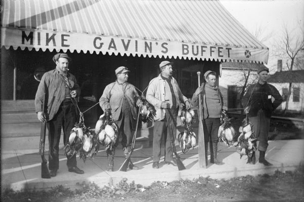 Five duck hunters stand in front of Mike Gavin's Buffet holding guns, paddles, and stringers of dead ducks.