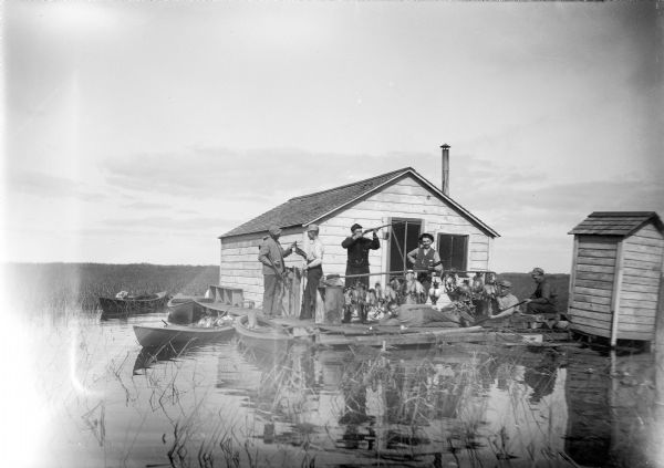 View across water towards a group of seven men posing on a dock in front of a small hunting shack in the middle of a marsh. Ducks hang on stringers attached to a wooden plank suspended along the dock. Duck hunting boats are tied to the dock on the left, and one boat has a pile of duck decoys. An outhouse is on the right edge of the dock, suspended over the water.