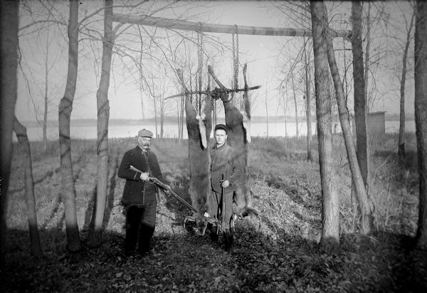 Two hunters pose in a wooded area. Their kill, two deer, hang upside down from a wooden beam supported by branches of trees. The deer have what appear to be tags on their antlers. Both hunters hold guns; one man stands between the deer, while the other man stands on the left. There is a lake or river in the background.