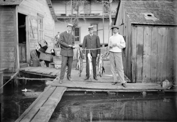 Three men (Korn brothers), all wearing hats, and two of them in suits, hold a bamboo pole holding stringers of fish between them. They are standing on a dock on the banks of Wolf River. Shacks and crates are behind them on the dock, and in the background a man stands near a building.