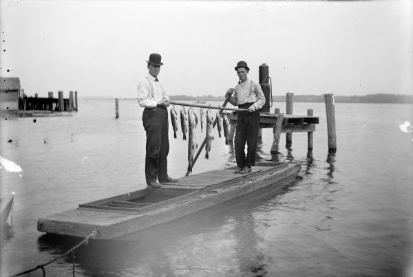 Two men stand on a flat boat on Wolf River. They hold stringers of fish suspended from a wooden pole between them. Behind them is what may be a gas pump on a platform supported by wooden pilings. In the background is the far shoreline. On the left is a building and a dock.