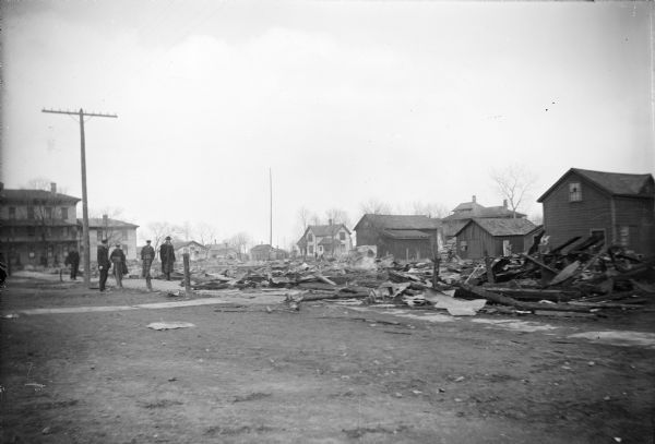 Five men standing on the far left survey fire damage on Main Street between N. 2nd and N. 3rd St in Winneconne. The area is completely destroyed. Surrounding houses and businesses unaffected by the fire are in the background.