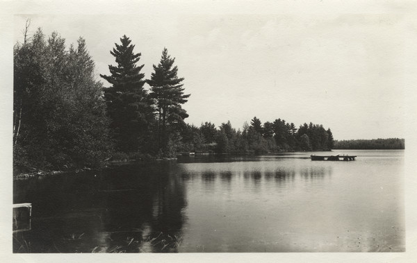 View from Camp Pioneer looking east along the north shore of bay (Lake Raymond, now known as Seventeen Lake). There are piers on the shoreline, and a raft on barrels with a diving board in the water. Original caption notes: "The shore line of the entire Lake is indented and heavily wooded. The Lake is sandy-bottomed, and there are no stumps or low places. Our typography [sic] is 100% for camp health, safety, and sanitation."
