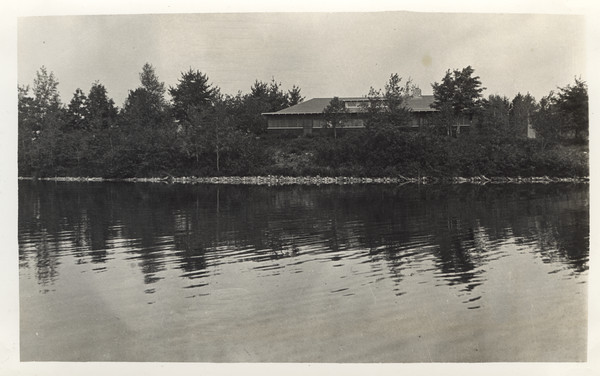 View over water of Main Lodge as seen from boat on bay. Original caption notes: "It fits well into the landscape and seems to have been there always. Forty years ago our site was an old lumber camp, which lends historical interest to it. The view from the porch, where we eat, out over the Lake is very inspiring and our campers enjoy it to the utmost."