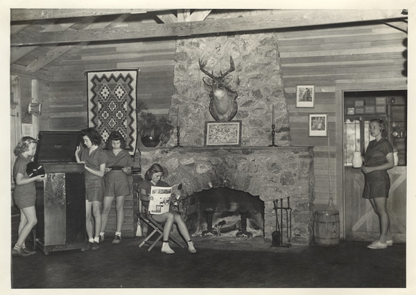 Interior view of stone fireplace area in the Main Lodge. Three campers are gathered around a record player, another is seated in a camp chair at the fireplace reading, and another is standing in a half-doorway holding a pitcher. The campers are all wearing the camp uniform. A deer head with antlers is mounted over the fireplace, and framed pictures and a woven rug hang on the wall. Original caption notes: "The stones for [the fireplace] were carefully selected by the builder, and the colours of the rocks, in sun or firelight, are very beautiful. On sunny days the fireplace does not attract the campers, but at night or on our few rainy days, it is quite a different matter. The door to the right leads to the kitchen, and from it half of the dining porch is served."