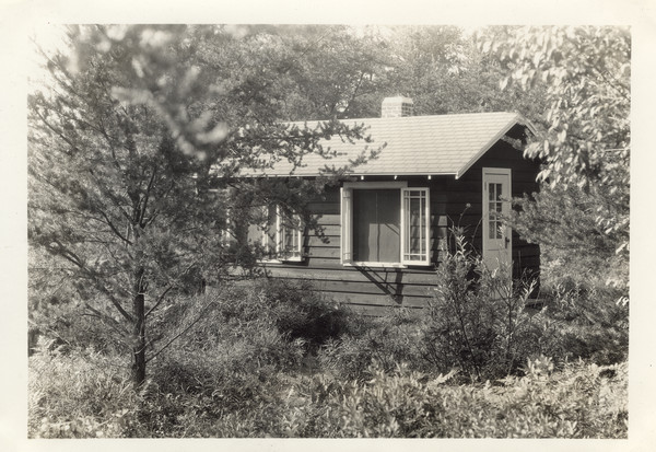 The health cabin located in a wooded setting. Original caption notes: "Here our medical counselor lives and keeps her equipment. In a separate room are two beds for people who need an extra nap or temporary rest. Our program is so arranged that each individual camper receives exercise and rest best suited for her own health improvement. Health benefits obtained at The Joy Camps last LONG after camping days are over!"