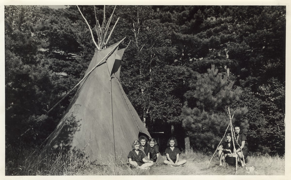 Exterior view of Indian tipi (teepee) with several campers sitting on the ground in front. Original caption notes: "One of the two attractive Indian tipis which we own. This one is on the shore front between the Workshop and the Directors' Cabin, and belongs to Deerpath Lodge. The Pioneer tipi is on our point across the Bay from Cabin III, and has a fine view down the Lake. These tipis are used a great deal for marshmallow roasts, for suppers, and for many other small group activities."