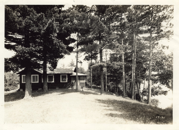 Exterior view of Directors' Cabin situated in wooded area, with a slope down to the lake on the right. Original caption notes that this cabin "also serves as the office and the general 'reception room' of our Camps. The campers use it constantly for special meetings, and often groups cook suppers over the fireplace. Sunday Vespers are held under the pines. All of our buildings are of the same attractive type of architecture and of the same substantial construction."
