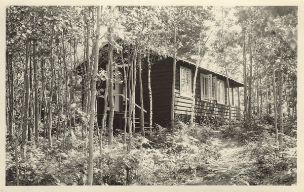 Exterior view of Cabin III situated in wooded area. Original caption notes that this was one of five "living cabins." The caption also notes: "These five living cabins are attractive, roomy, completely screened, comfortable, well-ventilated and dry in ALL sorts of north-woods weather. Each has a  suitable exposure, which affords adequate shade and sunlight. Each is placed advantageously on the camp site, with an attractive view on all sides. Each has a large porch, and a stove which is available for cold or wet weather. In each live three mature counselors and nine or ten girls, all of the same approximate age. Experience in group living in this sociologically sound set-up is an invaluable factor in the all-around development of the girl of today."