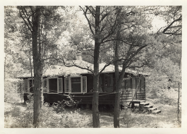 Exterior view of Cabin II situated in wooded area. Original caption notes that this was one of five "living cabins." The caption also notes: "These five living cabins are attractive, roomy, completely screened, comfortable, well-ventilated and dry in ALL sorts of north-woods weather. Each has a  suitable exposure, which affords adequate shade and sunlight. Each is placed advantageously on the camp site, with an attractive view on all sides. Each has a large porch, and a stove which is available for cold or wet weather. In each live three mature counselors and nine or ten girls, all of the same approximate age. Experience in group living in this sociologically sound set-up is an invaluable factor in the all-around development of the girl of today."