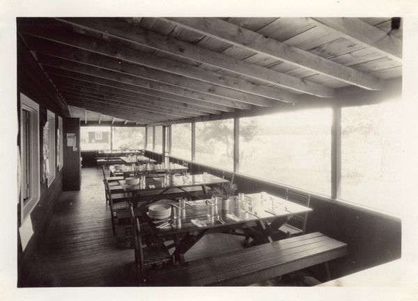 Interior view of the Dining Porch, which ran the entire length of the Main Lodge. View from porch is to the southeast, with a view of Lake Raymond (now known as Seventeen Lake). Original caption notes: "Our dining Porch runs the whole length of the Main Lodge, and enjoys a south-east exposure and a beautiful view of the Lake. Six girls and two counselors sit at each table. The furniture is all hand-made, and the silver and china attractive. Ask any old Joy Camper about the type, quantity, and attractiveness of the food we eat."