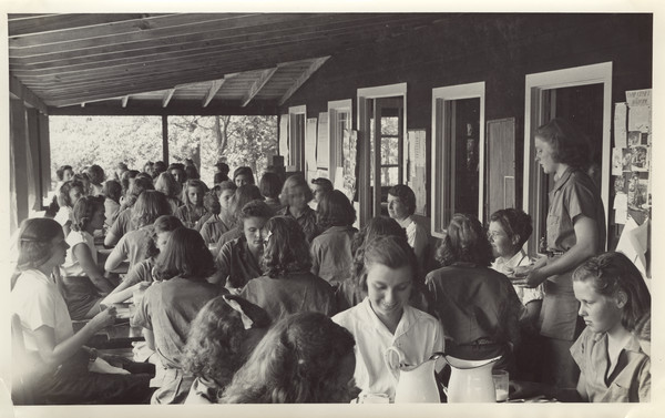 Interior view of campers and counselors eating on Dining Porch. Original caption notes: "Our dining Porch runs the whole length of the Main Lodge, and enjoys a south-east exposure and a beautiful view of the Lake. Six girls and two counselors sit at each table. The furniture is all hand-made, and the silver and china attractive. Ask any old Joy Camper about the type, quantity, and attractiveness of the food we eat."