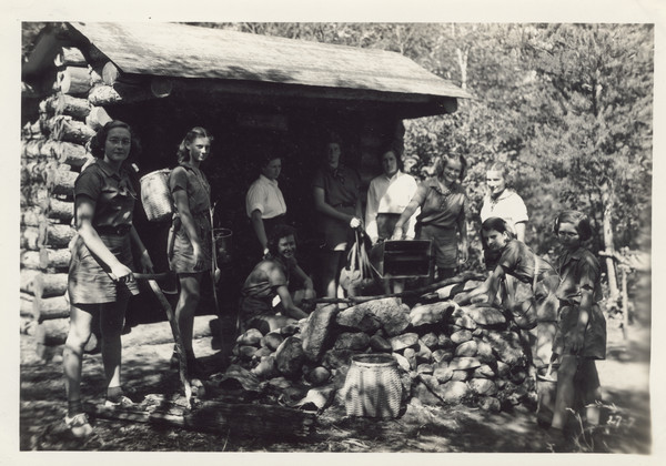 Exterior view of Adirondack Lean-to with several campers gathered around the stone fireplace. Some of the campers are holding or carrying an axe, a pocket knife, a pail, and a basket. Original caption notes: "Our Adirondack lean-to is situated across the Bay from the Main Lodge, and may be reached by trail and by water. With its deep floor of fragrant balsam boughs and with its large fireplace and fine view of the Lake, it is a favourite spot for outdoor meals, and overnight trips."