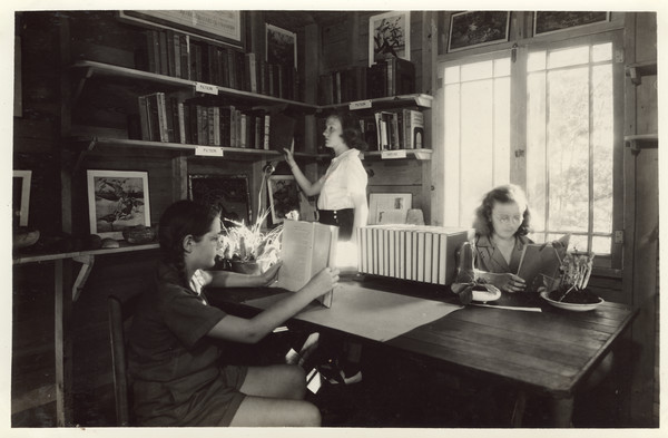 Interior view of library in Main Lodge. Two campers are seated at a table reading; another camper is browsing the shelves. Original caption notes: "In 1937, we established this Library in a pleasant corner of the Main Lodge. Each year the State of Wisconsin loans us a splendid collection which, added to the Camp library makes available to the campers several hundred volumes of fiction and stories, poetry, nature lore, and activity books. It is tastefully decorated with coloured pictures of game birds and Currier and Ives prints of outdoor subjects."