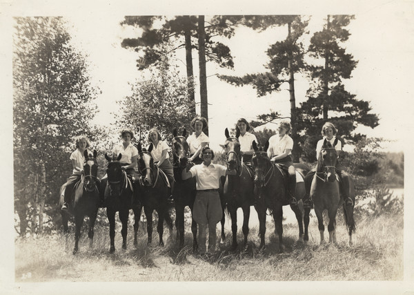 Group portrait of campers on horseback. One of the camp counselors is posed standing in the middle of the group. There is a lake is in the background. Original caption notes: "Riding is one of the most popular sports. English style of riding is taught, and complete at-home-ness on the horse is emphasized. The girls also learn how to saddle and bridle, and take general care of their mounts. Each horse is carefully selected, and all are three-gaited. We have a ring on the camp site, and hundreds of miles of woods trails available to us."