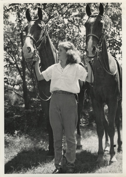 Full-length portrait of a young woman wearing jodhpurs standing and holding the reins of two horses. Original caption notes: "Riding is one of the most popular sports. English style of riding is taught, and complete at-home-ness on the horse is emphasized. The girls also learn how to saddle and bridle, and take general care of their mounts. Each horse is carefully selected, and all are three-gaited. We have a ring on the camp site, and hundreds of miles of woods trails available to us."
