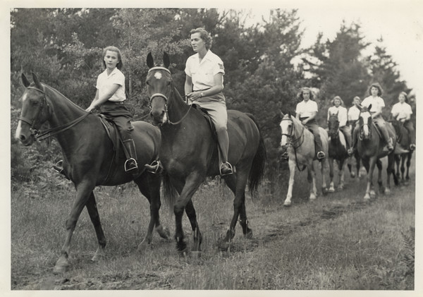 Group of campers and a camp counselor riding on horseback on a trail in wooded area. Original caption notes: "Riding is one of the most popular sports. English style of riding is taught, and complete at-home-ness on the horse is emphasized. The girls also learn how to saddle and bridle, and take general care of their mounts. Each horse is carefully selected, and all are three-gaited. We have a ring on the camp site, and hundreds of miles of woods trails available to us."