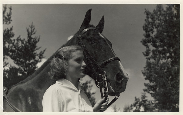 Portrait of young woman standing and holding the reins of a horse. Original caption notes: "Riding is one of the most popular sports. English style of riding is taught, and complete at-home-ness on the horse is emphasized. The girls also learn how to saddle and bridle, and take general care of their mounts. Each horse is carefully selected, and all are three-gaited. We have a ring on the camp site, and hundreds of miles of woods trails available to us."