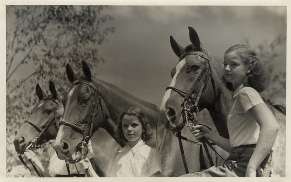 Group portrait of three young campers standing and holding the reins of three horses. Original caption notes: "Riding is one of the most popular sports. English style of riding is taught, and complete at-home-ness on the horse is emphasized. The girls also learn how to saddle and bridle, and take general care of their mounts. Each horse is carefully selected, and all are three-gaited. We have a ring on the camp site, and hundreds of miles of woods trails available to us."