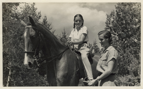 Camp counselor assisting with stirrups of a young camper sitting astride horse in a wooded area. Original caption notes: "Riding is one of the most popular sports. English style of riding is taught, and complete at-home-ness on the horse is emphasized. The girls also learn how to saddle and bridle, and take general care of their mounts. Each horse is carefully selected, and all are three-gaited. We have a ring on the camp site, and hundreds of miles of woods trails available to us."