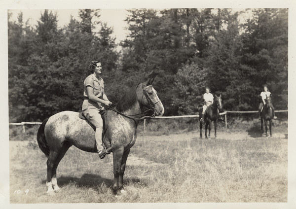 Camp counselor on horseback in a open field, with two campers on horseback in the background on the right near a fence. Trees are behind the fence. Original caption notes: "Riding is one of the most popular sports. English style of riding is taught, and complete at-home-ness on the horse is emphasized. The girls also learn how to saddle and bridle, and take general care of their mounts. Each horse is carefully selected, and all are three-gaited. We have a ring on the camp site, and hundreds of miles of woods trails available to us."