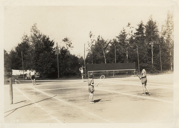 Doubles tennis match in play on tennis court in a wooded area. Original caption notes: "Our three most popular Land Sports are Tennis, Archery, and Baseball.... Every activity in camp is directed by a counselor especially trained, and the campers all realize and make the most of the opportunities which The Joy Camps offer for learning or improving on, all sports skills. Our equipment is excellent, instruction and cooperation of the VERY best."