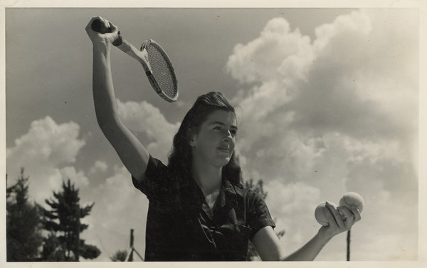 Camper poised with racket to serve tennis ball. Her racket is raised in the air, and she is holding two tennis balls in her left hand. Original caption notes: "Our three most popular Land Sports are Tennis, Archery, and Baseball.... Every activity in camp is directed by a counselor especially trained, and the campers all realize and make the most of the opportunities which The Joy Camps offer for learning or improving on, all sports skills. Our equipment is excellent, instruction and cooperation of the VERY best."