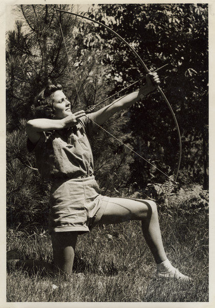 Profile view of archer kneeling in the grass on one knee, with bow and arrow ready and pointing towards the sky. Original caption notes: "Our three most popular Land Sports are Tennis, Archery, and Baseball.... Every activity in camp is directed by a counselor especially trained, and the campers all realize and make the most of the opportunities which The Joy Camps offer for learning or improving on, all sports skills. Our equipment is excellent, instruction and cooperation of the VERY best."