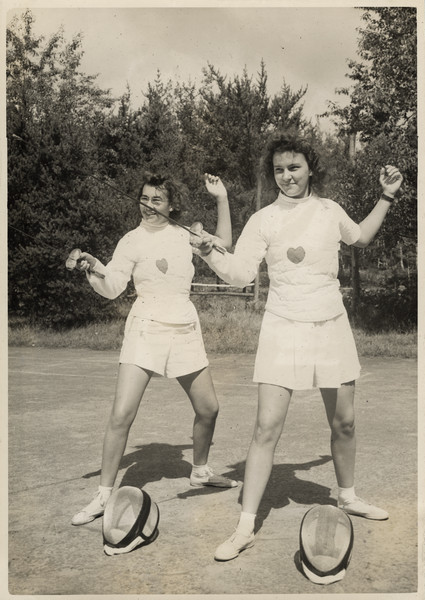 Two campers pose wearing fencing outfits and holding epees, with protective headgear sitting on the ground. They are standing on a court or concrete pad. Original caption notes that fencing was added as a sport to camp activities in 1941, and it quickly became very popular with the girls.