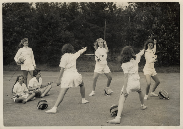 Two sets of fencing partners are posed in fencing stance, holding epees, and wearing protective clothing. Their headgear is sitting on the court near their feet. Three other campers are on the left watching. Original caption notes that fencing was added as a sport to camp activities in 1941, and it quickly became very popular with the girls.