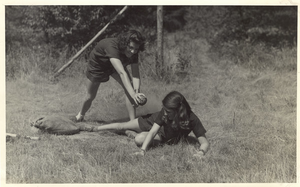 Two campers playing baseball, with one girl sliding into base and the other girl holding the ball. Original caption notes: "Our three most popular Land Sports are Tennis, Archery, and Baseball.... Every activity in camp is directed by a counselor especially trained, and the campers all realize and make the most of the opportunities which The Joy Camps offer for learning or improving on, all sports skills. Our equipment is excellent, instruction and cooperation of the VERY best."