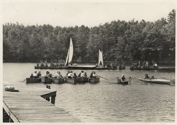 View from shoreline from pier of campers in boats on the water. Rowing crews and kayaks are in the foreground. Original caption notes that this image offers a sampling of the many different kinds of boats the girls learned how to use: rowboats, kayaks, including the Folbot variety; sailboats, paddle boats, canoes, including an Indian birch bark canoe (many of these are in the background). There is a diving board on the end of the pier. The caption also notes: "...with justified pride we can boast of the excellent instruction and the resulting careful preparation for participation in all water sports made possible by our outstanding corps of waterfront counselors. Our water sports program, under the personal direction of Miss Camp, is recognized as one of the best in camps for girls in this country and Canada."