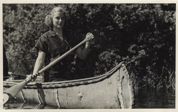 Young woman in birch bark canoe on a lake close to the shoreline. She is holding a paddle.