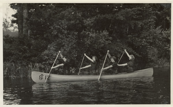 Group portrait of four young campers posed near the shoreline in a Joy Camps canoe displaying the Joy Camps logo (boat displays lettering and numbering "JC 12"), with oars poised in the water.