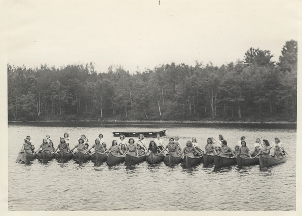 Large group portrait of campers and counselors sitting in a row of fourteen Joy Camps canoes, side-by-side in a lake. A raft floats in the water behind them.