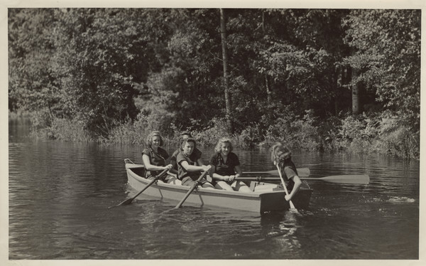 View across water of five young women in a rowboat near the shoreline.