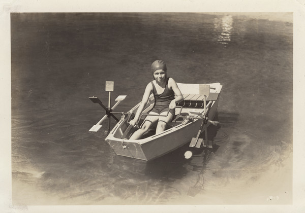 Young girl sitting in a paddleboat on the water. The girl wears a swimming cap and a one-piece swimsuit, and has her hands on the handles that spin the paddles.