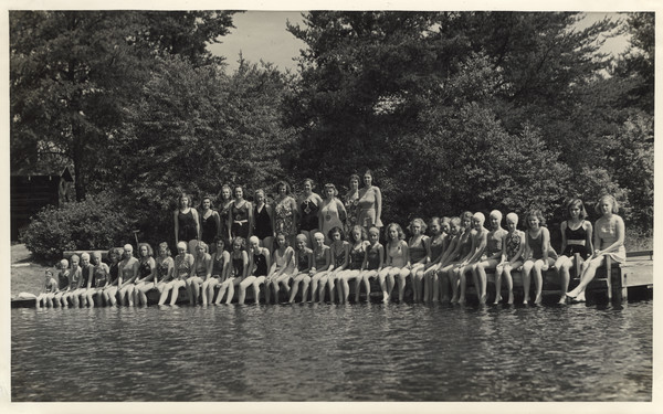 Large group portrait over water towards campers and counselors posed in two rows along a pier. They are all wearing swimming suits, and some wear swimming caps. The campers are seated on the dock with their counselors standing behind them. There is a cabin on the left among trees. Original caption notes: "Swimming in the clear, cool, PURE waters of our Lake Raymond is a thrill and a pleasure never to be forgotten. Diving, life-saving and water safety, and the harder strokes appeal to good swimmers. Beginners progress beyond the fondest dreams of their parents and of themselves. Long hours of training and experience stand behind those who staff our waterfront. Parents need never worry about their daughter's safety in our care."