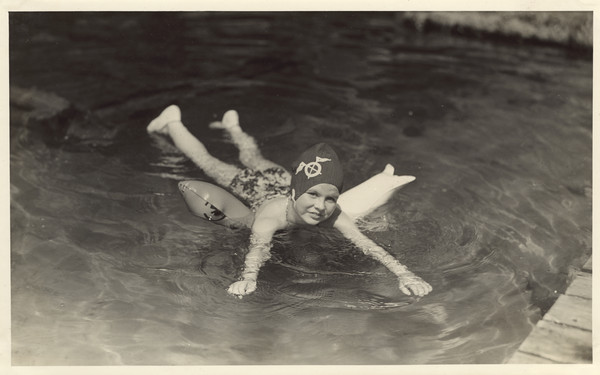 Young girl in bathing suit and swim cap floating in the water atop an inflated, plastic floating device. A wood pier is in the right foreground. Original caption notes: "Swimming in the clear, cool, PURE waters of our Lake Raymond is a thrill and a pleasure never to be forgotten. Diving, life-saving and water safety, and the harder strokes appeal to good swimmers. Beginners progress beyond the fondest dreams of their parents and of themselves. Long hours of training and experience stand behind those who staff our waterfront. Parents need never worry about their daughter's safety in our care."