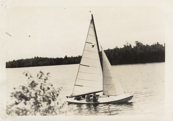 View from shoreline of sailboat on a lake, with several campers in the boat (mostly blocked from view by larger sail). The far shoreline is in the background. Original caption notes: "Sailing was added to our list of water activities in 1933, and it has been exceptionally popular. Our boat is a Snipe, with Genoa jib. Although safe, it is a sporty little boat, and campers who wish may learn all the details of handling a boat of this type. Needless to say, the Snipe is at its moorings only at mealtimes!"