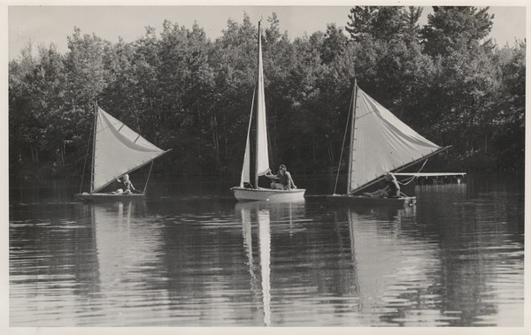 Exterior view of several campers handling three sailboats on a lake. Reflection of the sails are captured on the surface of water. The far shoreline is in the background. There is a raft floating in the water behind the sailboat on the right. Original caption notes: "Sailing was added to our list of water activities in 1933, and it has been exceptionally popular. Our boat is a Snipe, with Genoa jib. Although safe, it is a sporty little boat, and campers who wish may learn all the details of handling a boat of this type. Needless to say, the Snipe is at its moorings only at mealtimes!"
