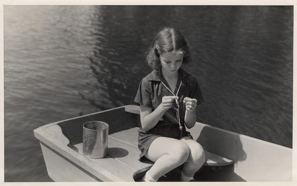 Young girl sitting in a rowboat on the water baiting a fishing hook. A metal can sits beside her on the seat. Original caption notes: "Perch, large and small mouth black bass, and other pan fish abound in our Lake. Our campers spend hours on end at this sport, and they love it. Fishing breakfasts add to the pleasure. And usually once a week we catch enough to feed our camp family of seventy-five, and that is not a fish-story, either!"