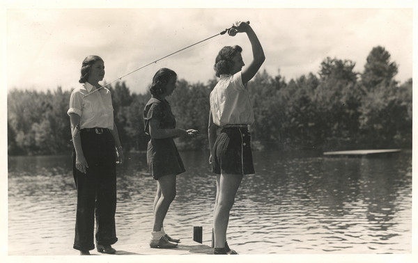 Three young women standing on a dock. The woman on the right has a fishing pole raised above her head and is ready to cast. There is a raft floating out on the lake, and the far shoreline is in the background. Original caption notes: "Perch, large and small mouth black bass, and other pan fish abound in our Lake. Our campers spend hours on end at this sport, and they love it. Fishing breakfasts add to the pleasure. And usually once a week we catch enough to feed our camp family of seventy-five, and that is not a fish-story, either!"
