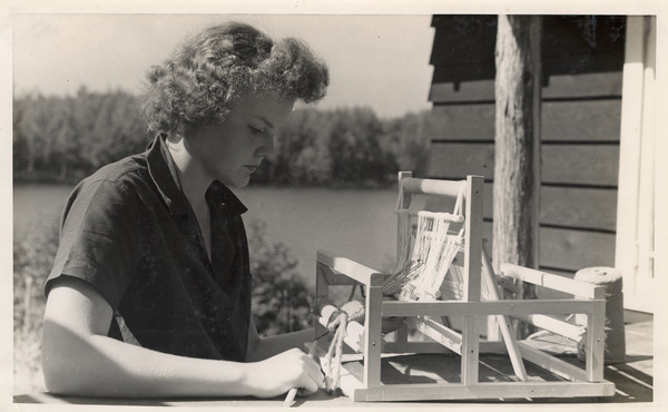 Close-up of young woman weaving with a table loom. She is sitting at a table outdoors next to a building. A lake and shoreline are in the background.