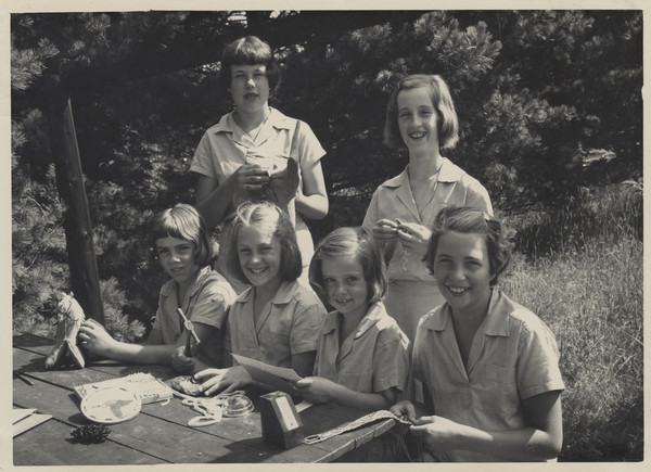 Outdoor group portrait of four campers seated at a wooden table and two campers standing behind them. They are working on camp arts and crafts projects.