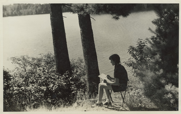 Young camper seated on stool on a hill overlooking the lake. She has a sketchpad and pencil in hand.
